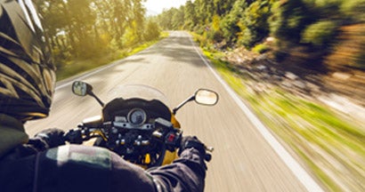 Person riding a motorcycle on a winding road through a sunlit, tree-lined landscape. Find Gap Motorcycle Insurance.