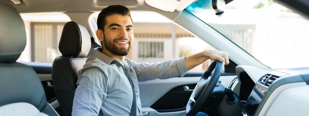 Hispanic man looking at the camera while driving his new car with pride. Cumberland Hill, Rhode Island Car Insurance. 