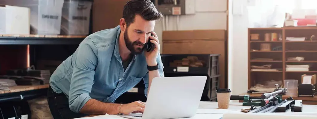Businessman using phone while designing a blueprint in a creative office. Find Shelburne, Vermont business insurance.