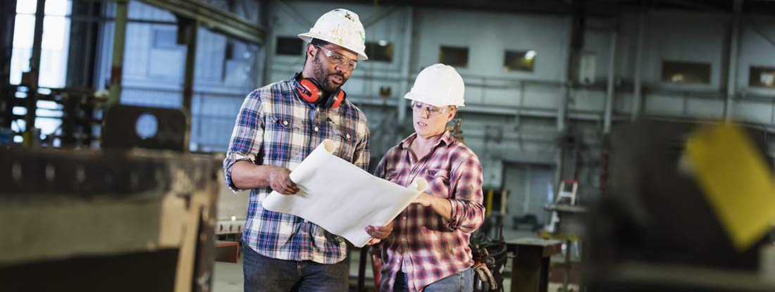 Man and woman standing on factory floor wearing hardhats and safety goggles, reading a set of plans. Self insured companies.
