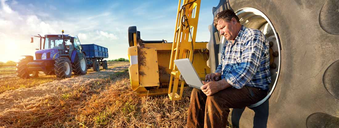 Farmer using a laptop in a field next to a tractor and other farm equipment. How much does farm insurance cost.