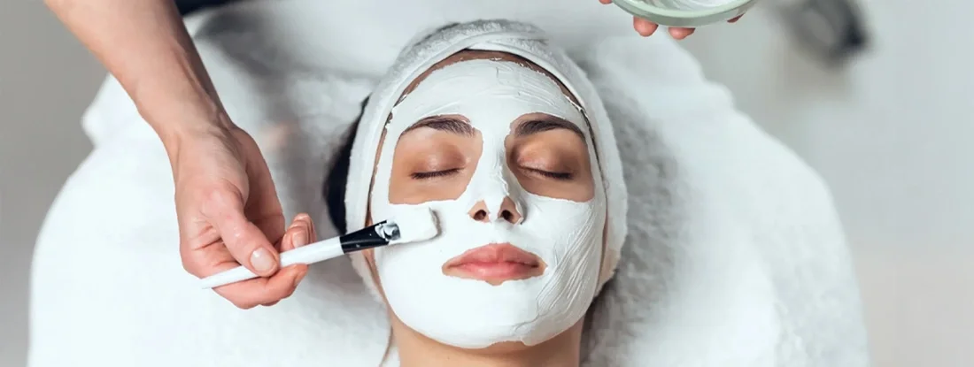 Cosmetologist applying facial mask to woman while lying on a stretcher in the spa center. Find Esthetician Insurance.