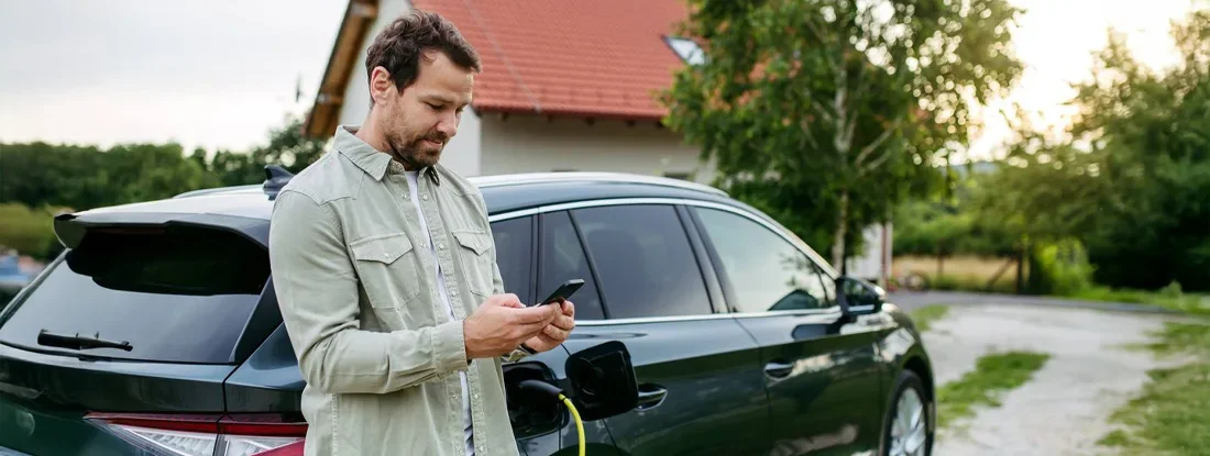 Man charging electric car in front of house, holding smartphone. How to Buy a Great Used Hybrid Car.