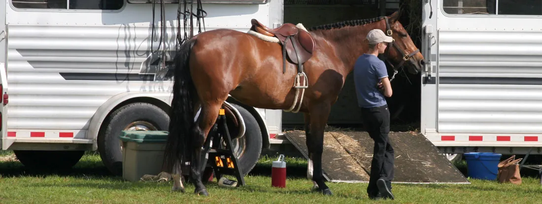 A horse and rider stand outside a large horse trailer in a grassy field. Find West Virginia Trailer Insurance.