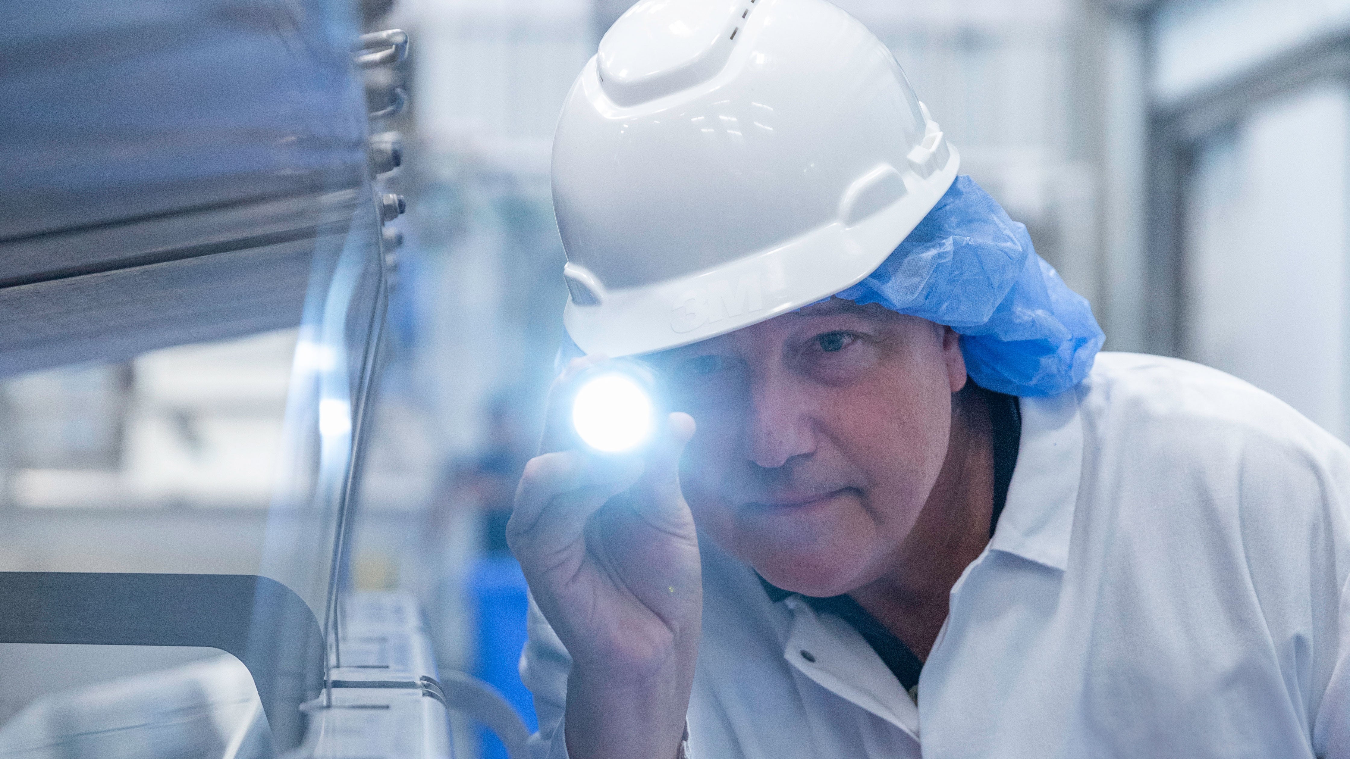 Um trabalhador com capacete branco e equipamento de proteção inspeciona o maquinário com uma lanterna em uma fábrica de processamento de alimentos