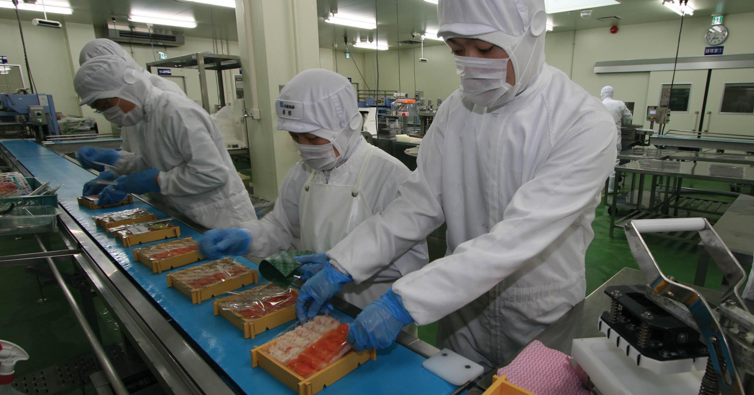 Several Sato Suisan employees in white uniforms with hygienic masks and caps seal bento boxes traveling along a ThermoDrive belt