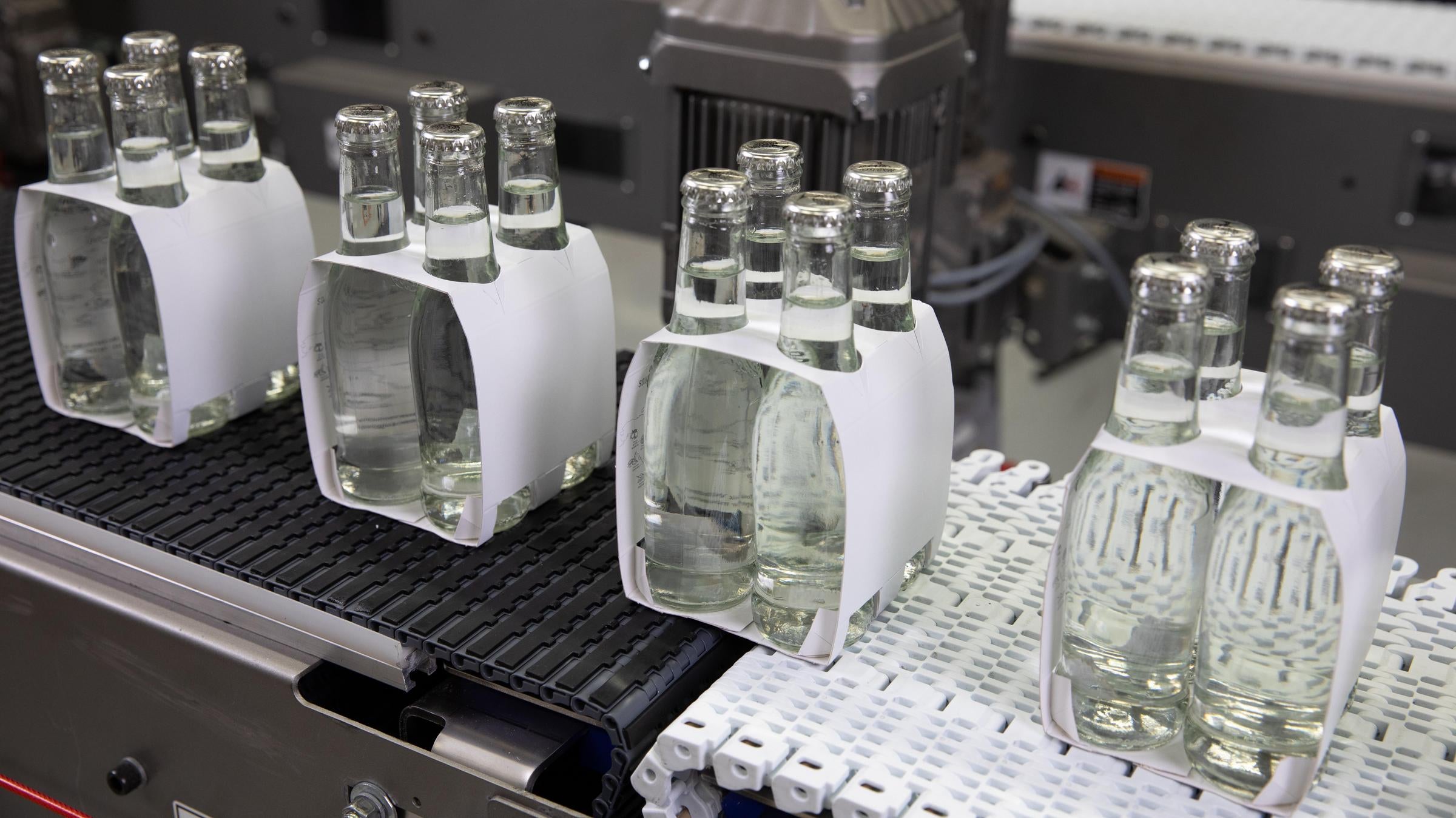 4 Packungen mit transparenten Flaschen mit klarer Flüssigkeit werden zwischen zwei Förderbändern übergeben