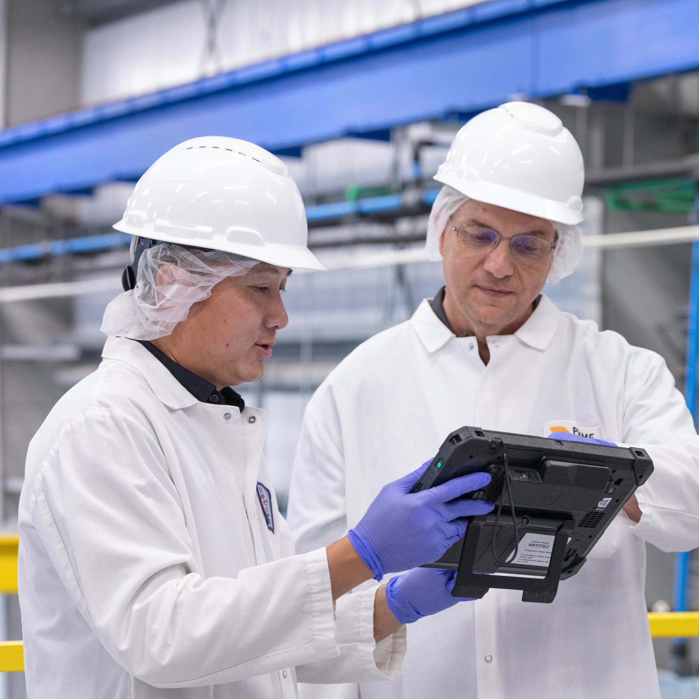 Dois homens com jalecos brancos e capacetes analisando um tablet eletrônico, juntos em uma fábrica