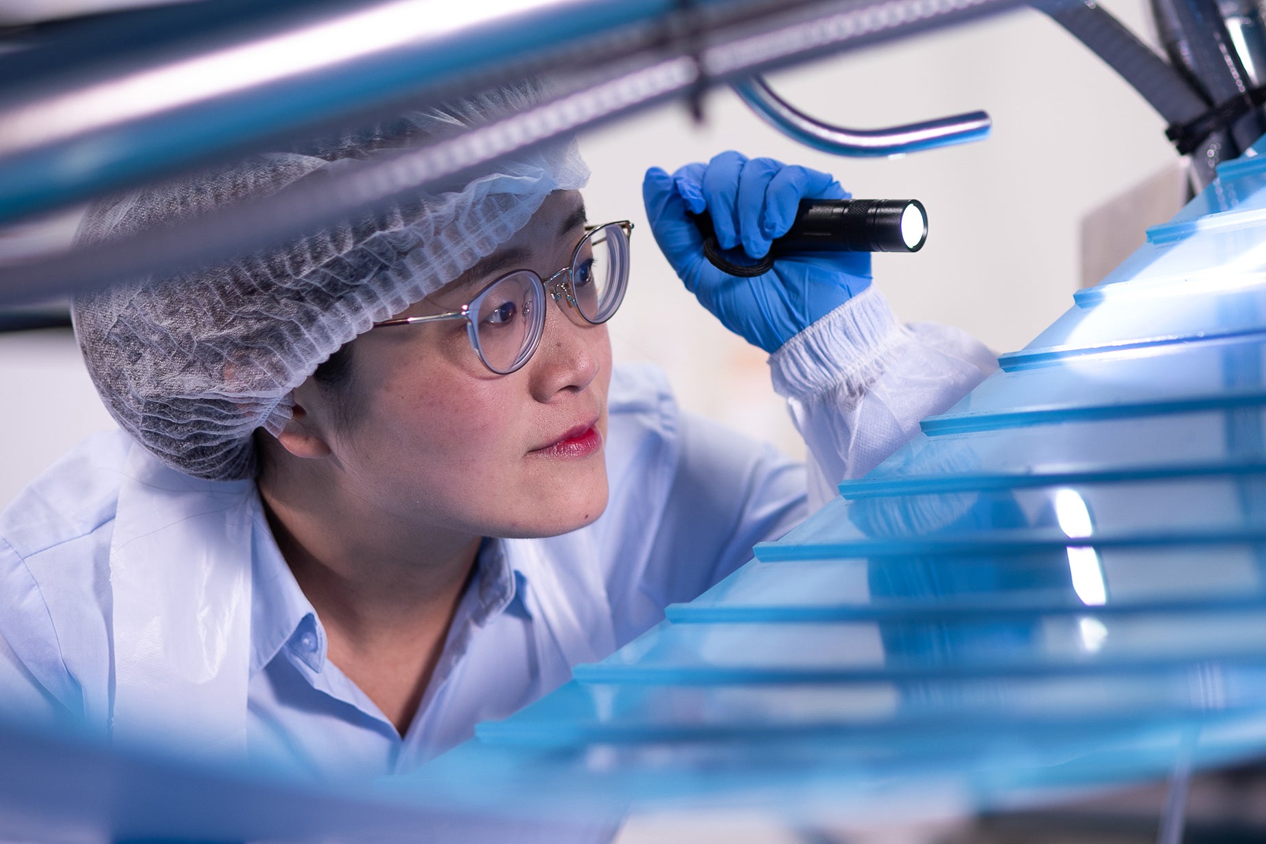 Trabalhadora com óculos, rede de cabelo e outros equipamentos de proteção usando uma lanterna para inspecionar a parte inferior da esteira transportadora ThermoDrive