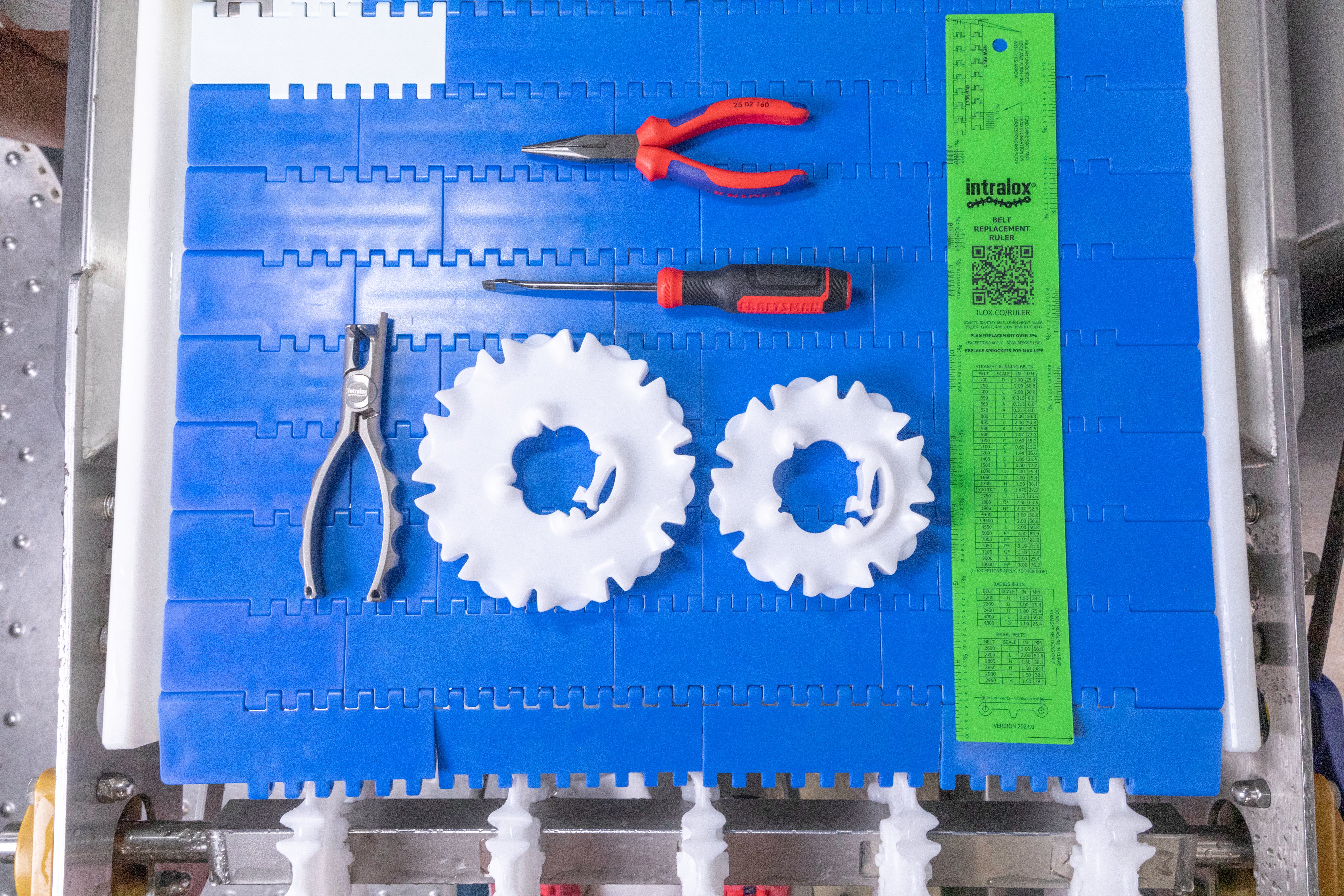 Top-down view of a blue modular plastic conveyor belt with various tools neatly arranged on it. Items include two white plastic sprockets, a pair of pliers, needle-nose pliers with red handles, a screwdriver, and a bright green Intralox belt replacement ruler. 