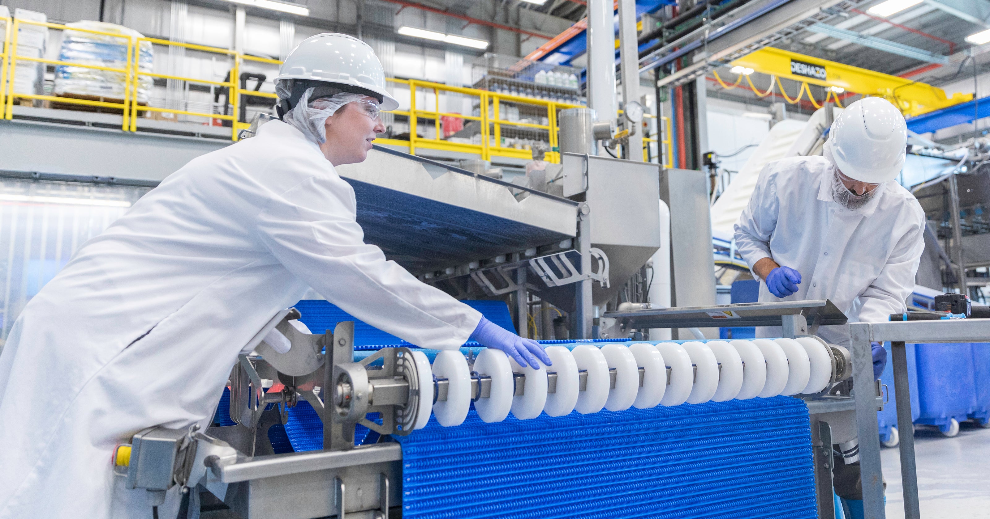 製造工場で青いFoodSafeコンベアベルトとホイールスプロケットを検査する、白衣と保護具を身に着けた2人の作業員