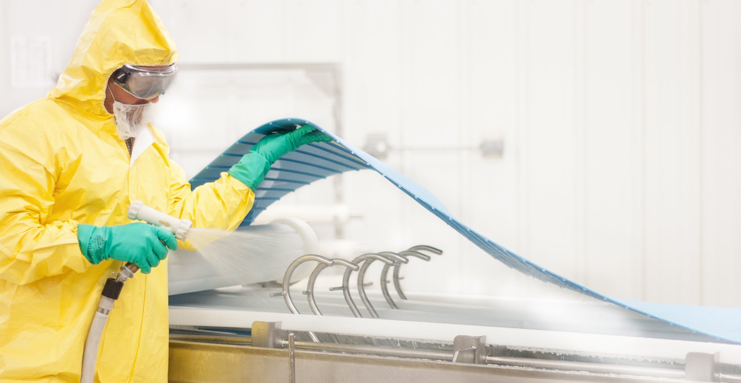 Homem vestindo equipamento de proteção amarelo completo, incluindo capuz, máscara e luvas, usando uma mangueira para pulverizar água na parte inferior de uma esteira transportadora ThermoDrive e de seu transportador