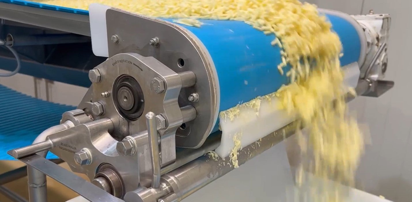 Close-up of shredded cheese moving on a blue conveyor belt in a stainless steel food processing machine, falling off the end in a steady stream.