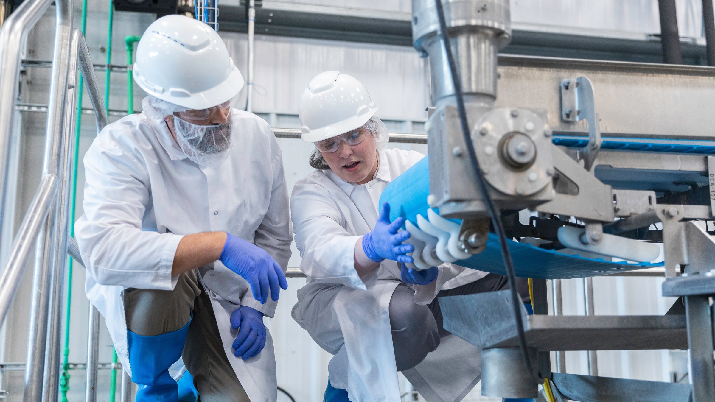 白いヘルメット、手袋、白衣などの安全衛生保護装備を身につけて工場でコンベアの検査を行っている男性1名、女性1名、合わせて2名の食品産業のプロフェッショナル。