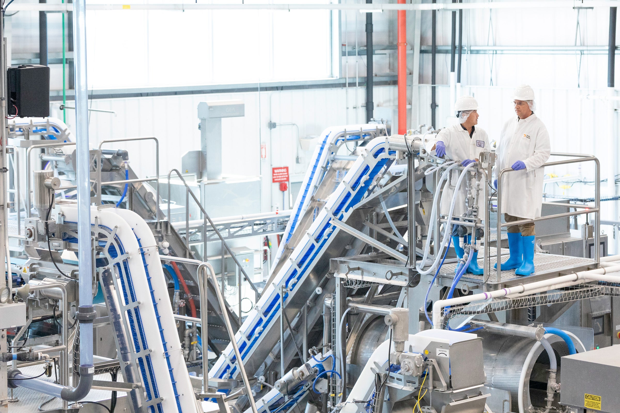 两位穿戴防护装备的操作人员站在工厂中输送机之间的平台上