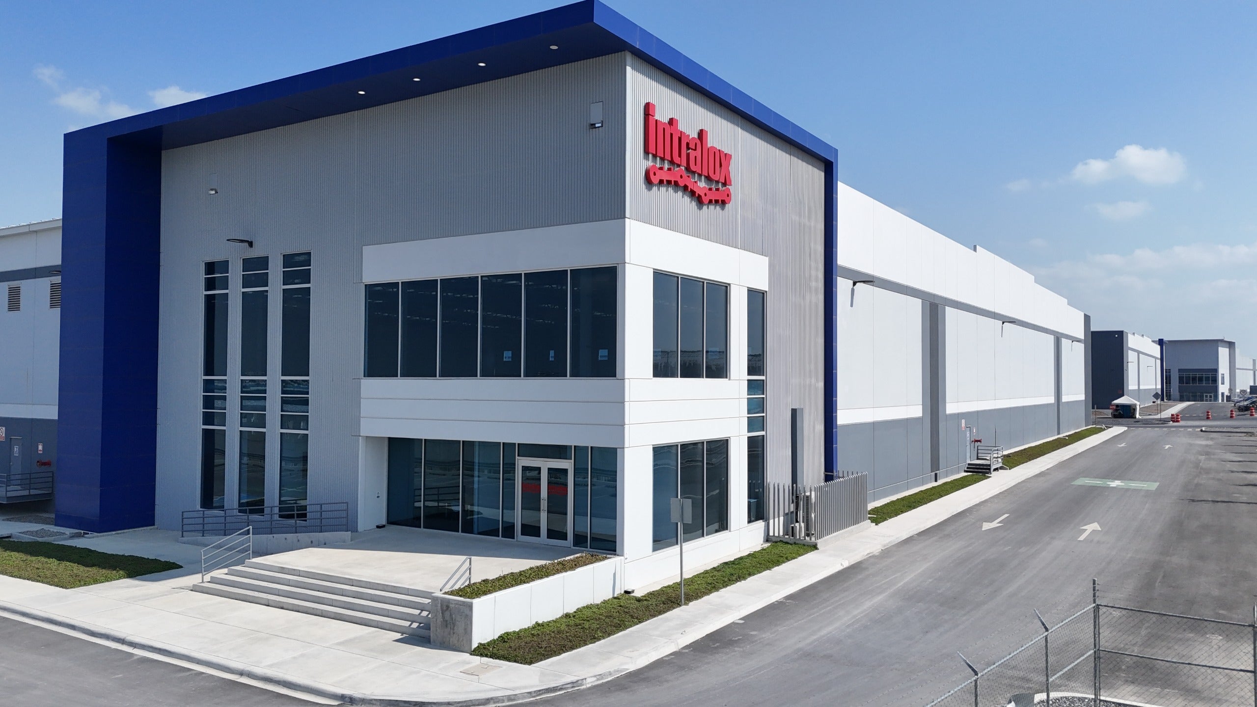 Fachada da nova unidade da Intralox no México, mostrando uma moderna entrada de vidro de dois andares com o logotipo Intralox e espaço industrial adjacente para armazenamento.