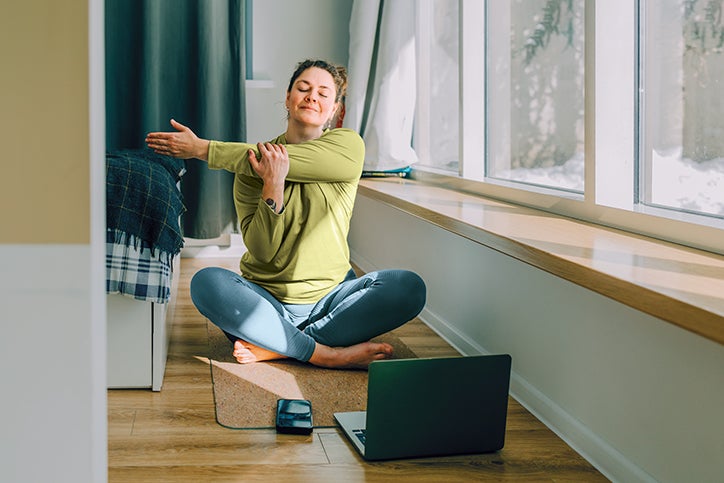 Lady sitting on the floor, in front of her laptop stretching.