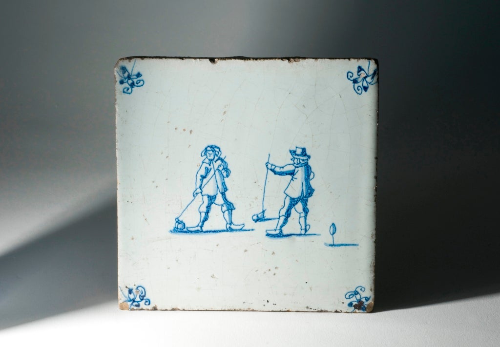 White, square ceramic tile decorated with blue figures in the middle. The two figures hold sticks to the floor with balls. The tile has four blue embellishments in each corner. 