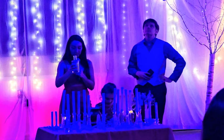 Two ritual leaders stand before a backdrop of fairy lights and white curtains, behind a table of candles.