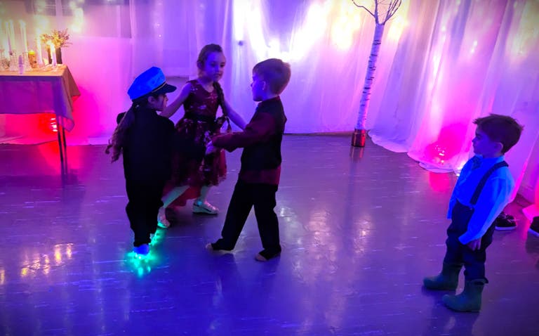 Young children dance together on the dance floor beside white birch branches and glowing draped curtains.