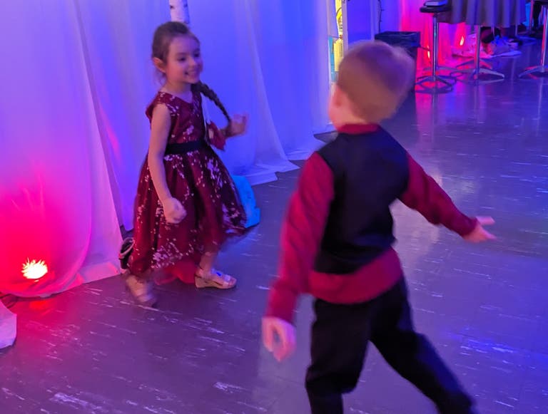 A girl in a sparkly red dress and a boy in a red shirt and black vest dance across the floor.