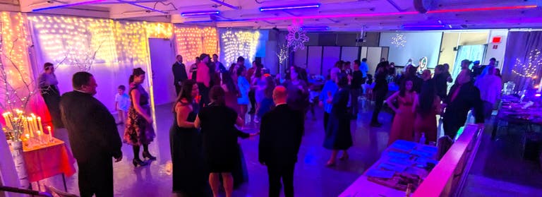 A crowded hall of guests in formal attire mingling and dancing beneath curtains of white fairy lights.