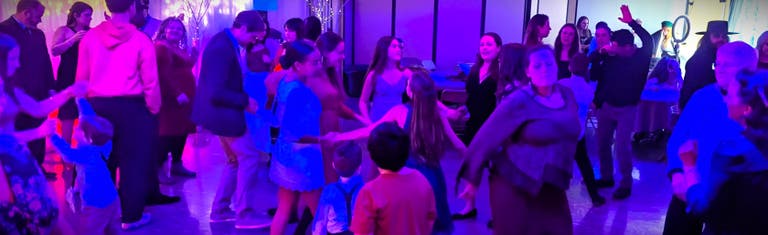 A packed dance floor of adults and children dancing under blue and purple lights.