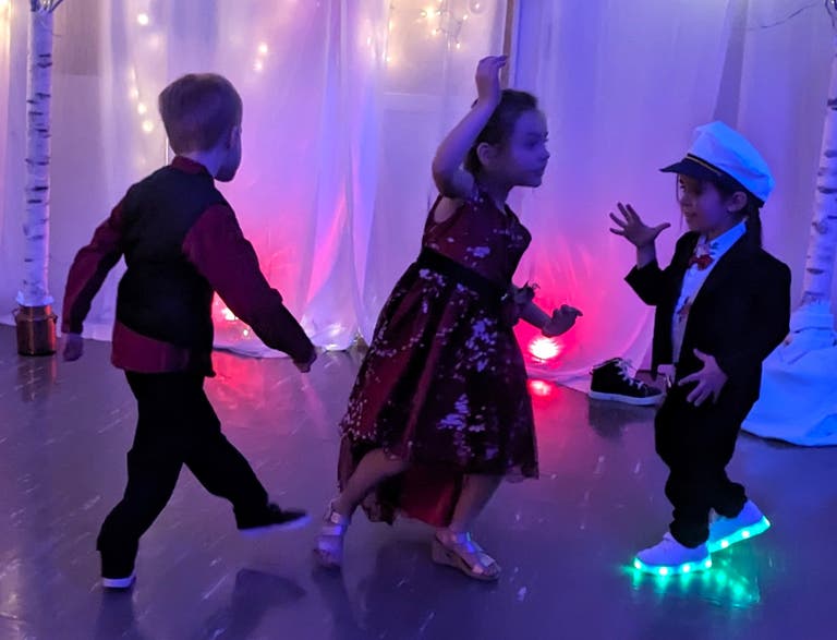 Three young children in formal outfits dance under purple lights, one in light-up sneakers and a white captain's hat.