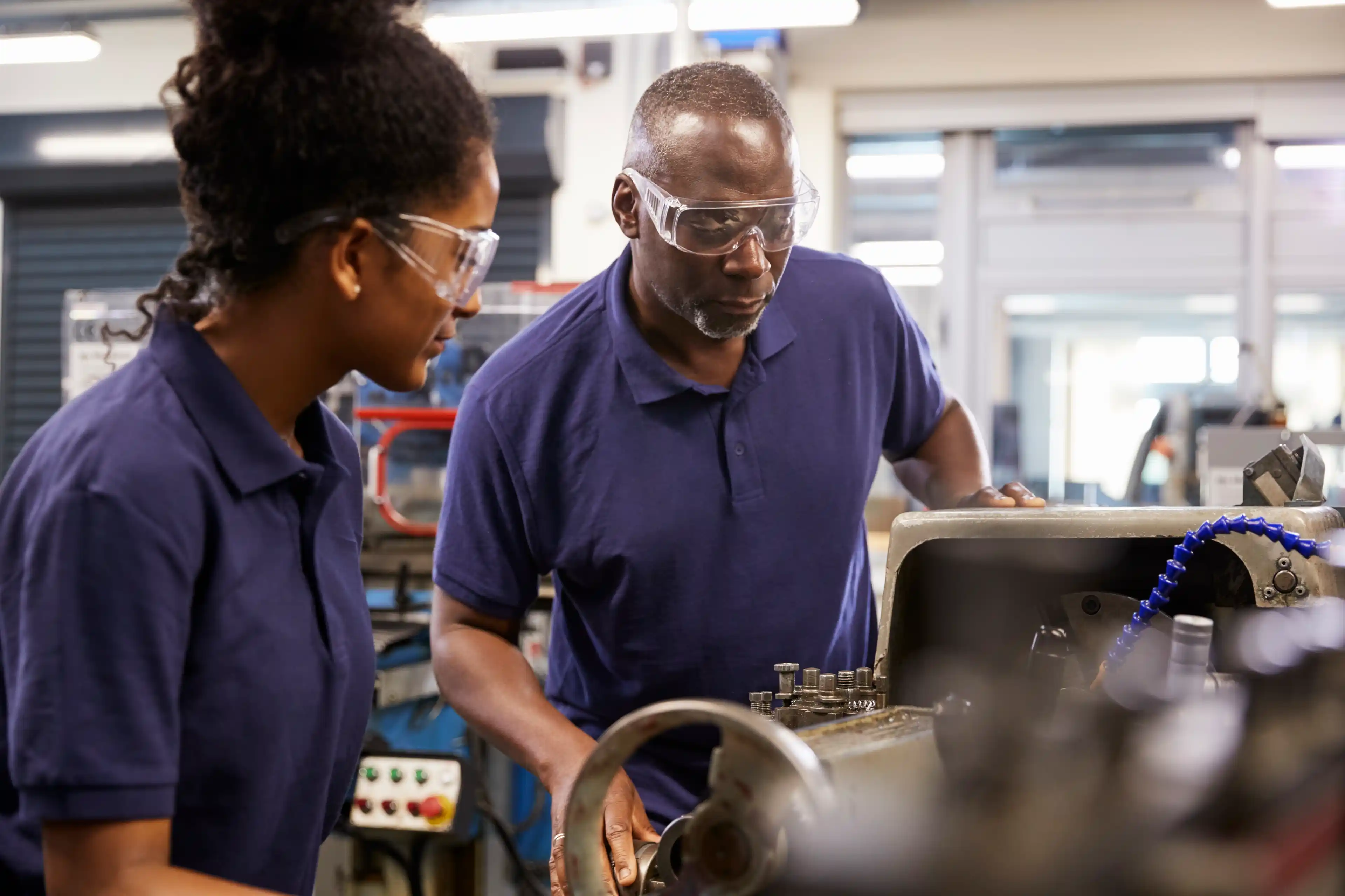 2 people in blue t-shirts and safety goggles working with a machine.
