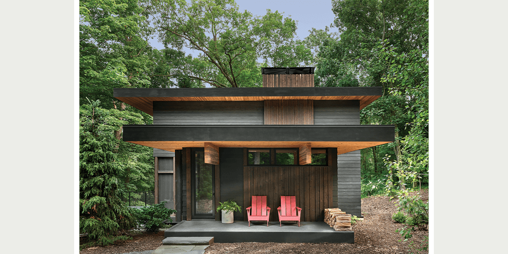 A front porch of a modern rustic home featuring two red Adirondack chairs amid a lush forest setting. 