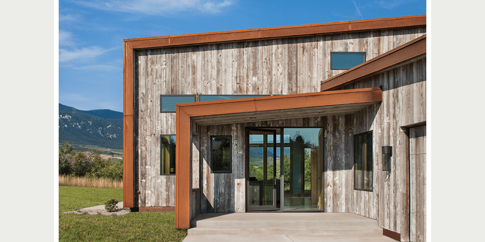 A modern rustic home features a wood exterior complemented by a glass swinging door with sidelite sheltered under a sloped roof.