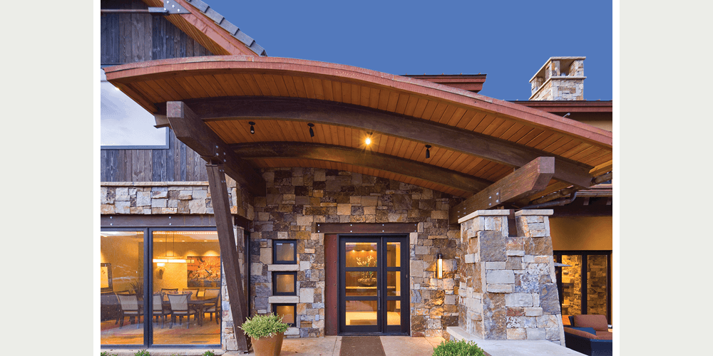 A modern mountain lodge-style home features a stone exterior with a sloped roof overhanging the entrance.