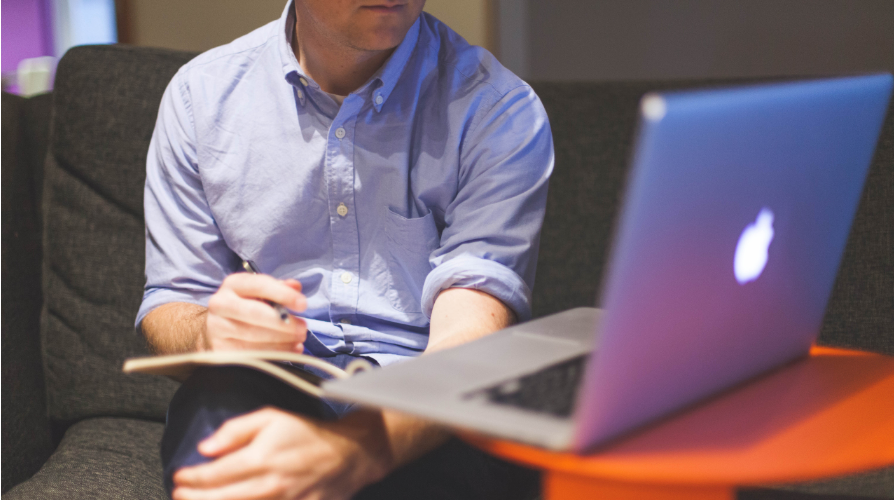 photo of man in blue shirt sitting on couch with book on his lap looking at macbook