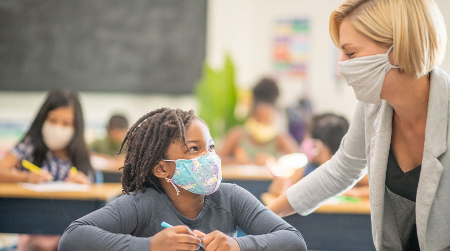 A young student with a mask on interacting with his teacher in a classroom