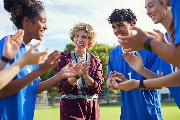 Coach and high school students clap in a huddle on an athletics field.