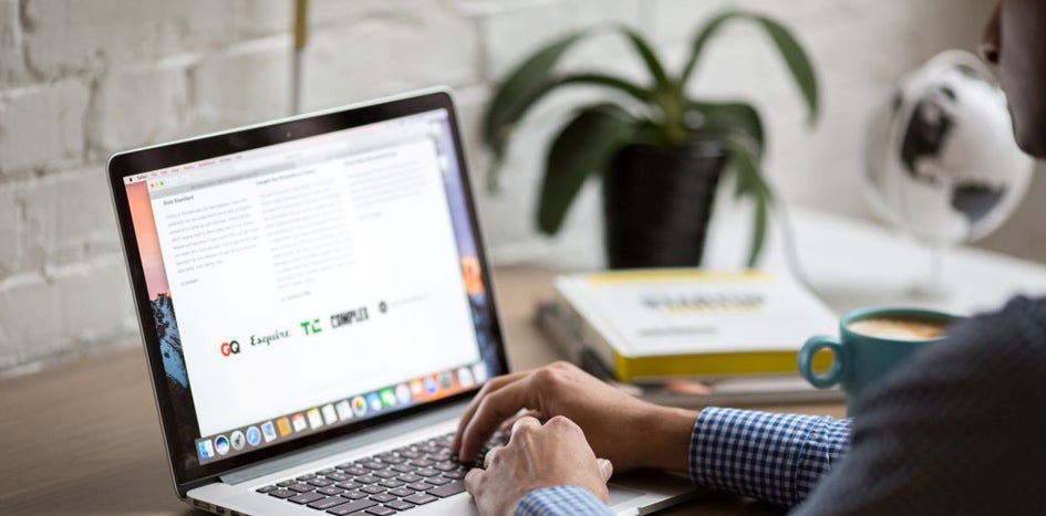 Man typing on a laptop on a desk with books and a plant on it.