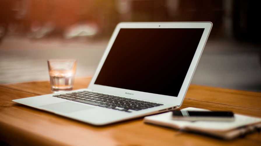 photo of a laptop on a desk placed between a glass of water and a notebook with a pen