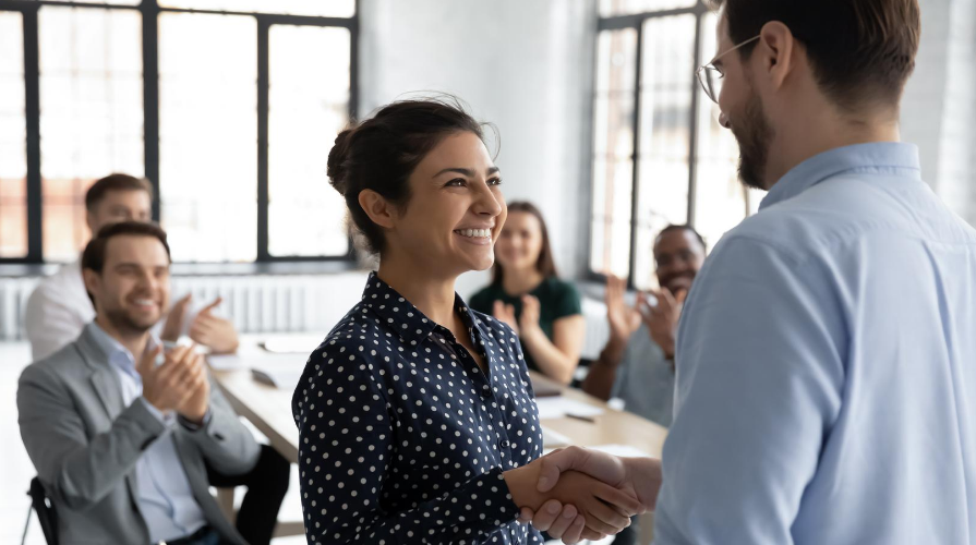 photo of a woman shaking hands with a man, co-workers applauding in the background