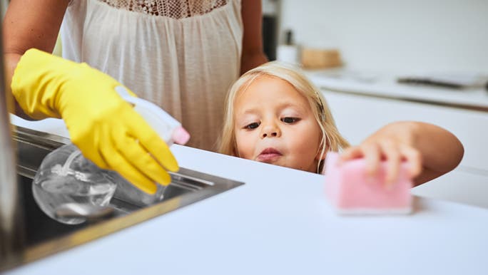 A little girl helps her mum wipe down the benchtop with a pink sponge. She is learning responsibility and building her resilience. 