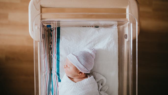 From above, a newborn is swaddled in a hospital bassinet. They have a cute beanie on and are sleeping soundly. 