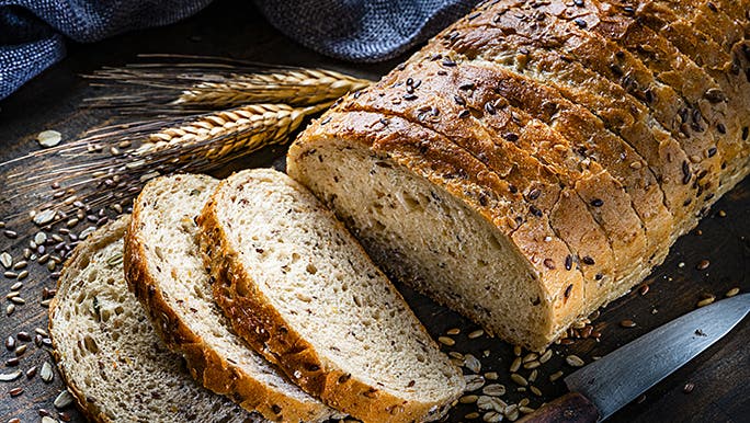 A loaf of multigrain bread, sliced on a chopping board, surrounded by grains and seeds.
