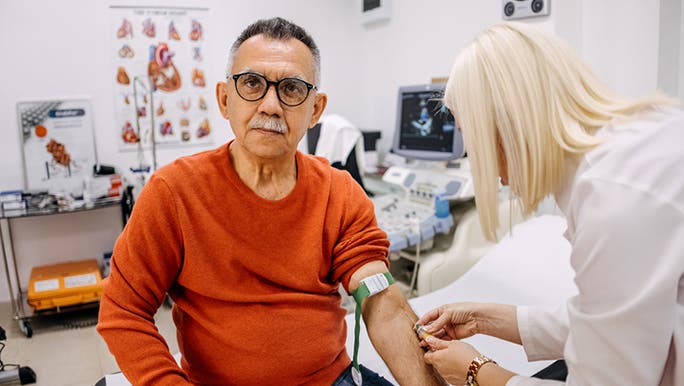 An olive-skinned man with dark hair and glasses wearing a red jumper is having a blood test