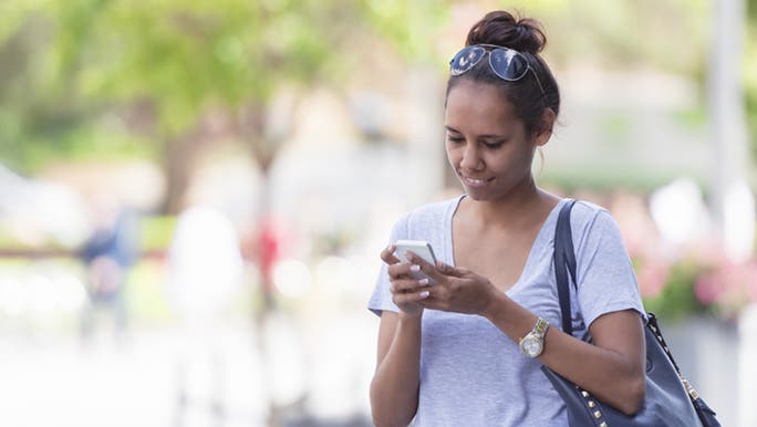 A woman stops during her walking commute to reply to a text message, she is happy and may be talking to a long distance friend. 