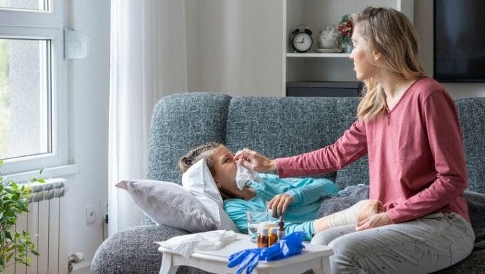 A mother is taking care of a sick child who is lying on a sofa, with bottles of pills on a nearby table