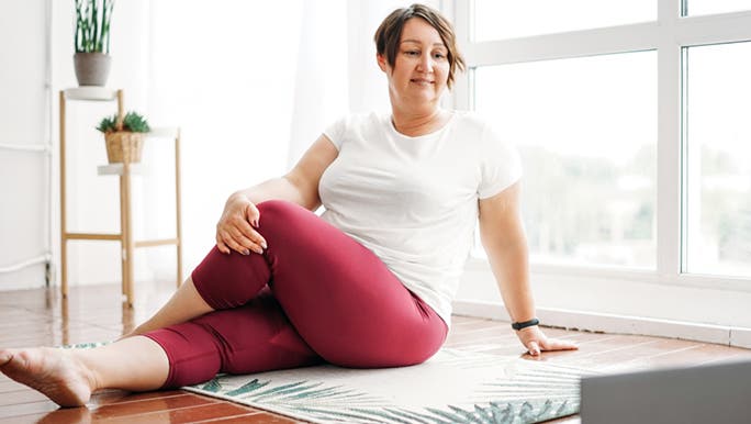 A woman stretches on a mat by a sunny window watching a video on her tablet about why women are more flexible.