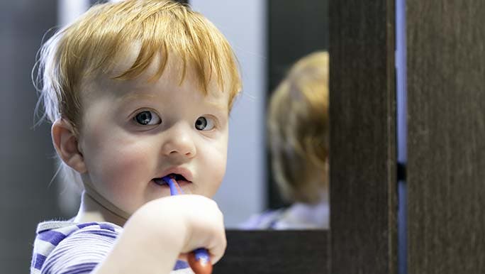 Toddler brushing his teeth at the mirror while his parents wonder how long it takes for baby teeth to grow