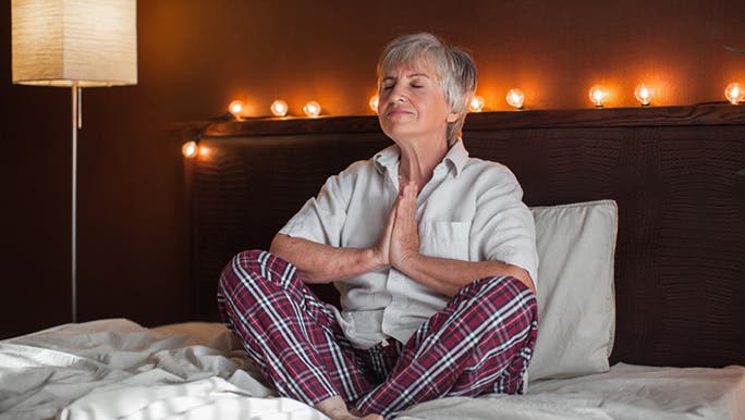 A senior woman wearing checked pyjamas is sitting in bed meditating before going to sleep
