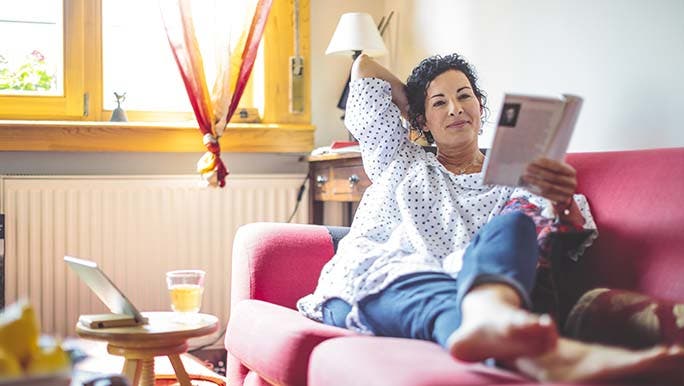 A woman lies on a bright pink couch, smiling while reading a self-care magazine.