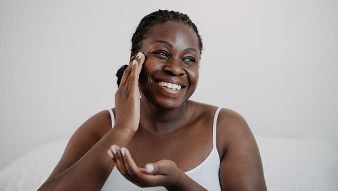 A woman in a white singlet applies cream to her face while smiling as she wonders if collagen powder is the same as gelatin.