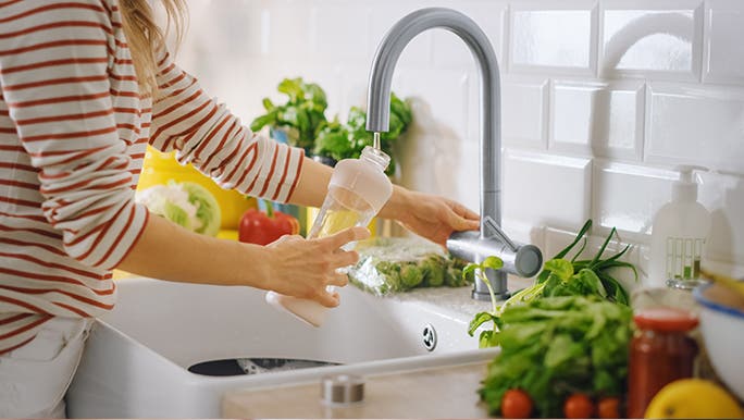 In a modern kitchen, a woman fills a drink bottle from the tap. The counter is packed with fresh fruit and vegetables. She is aiming to increase good bacteria in her gut naturally. 